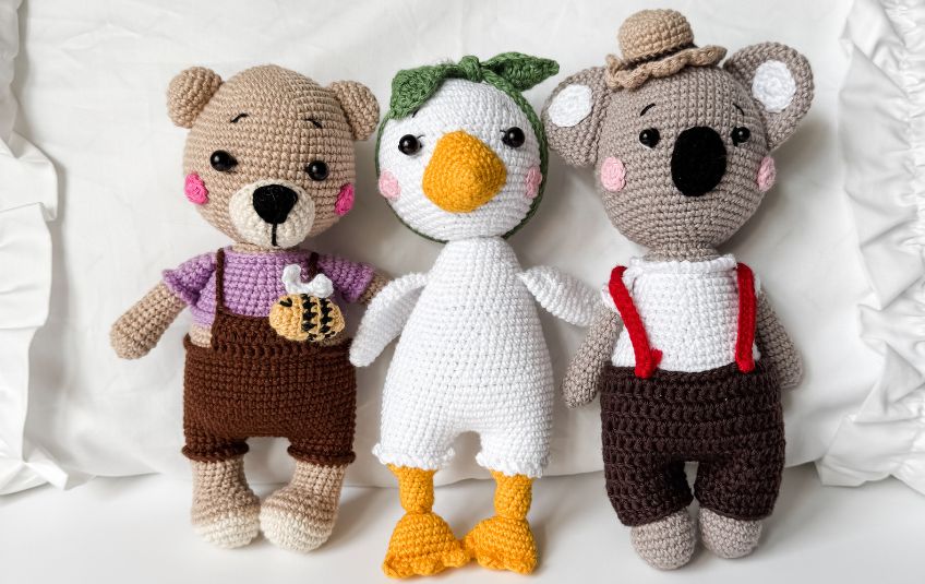Three amigurumi yarn handcrafted stuffed animals, bear with a bee, duck with a bow, koala with suspenders and a hat